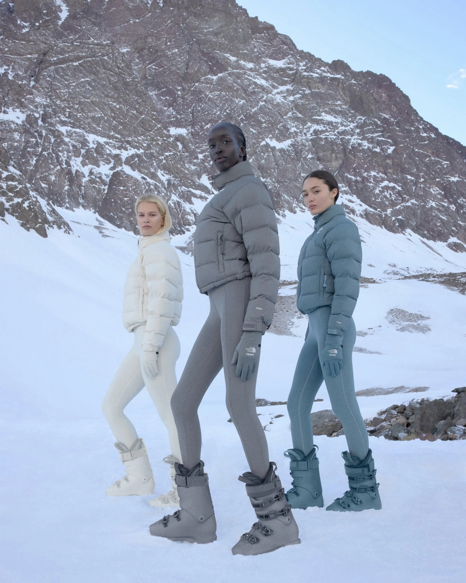 Three models in colorful The North Face x Skims down jackets and snow boots, walking on snow in front of a mountain