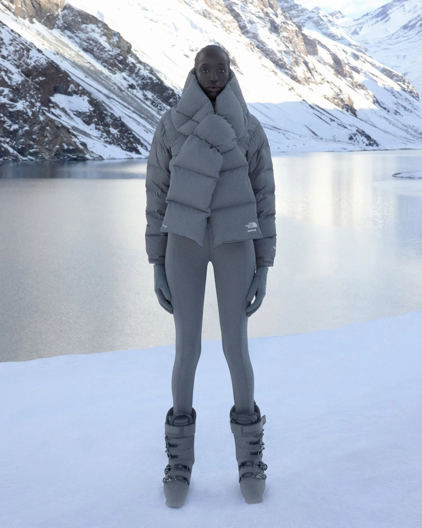 Modèle en tenue The North Face x Skims grise avec bottes, devant lac alpin enneigé