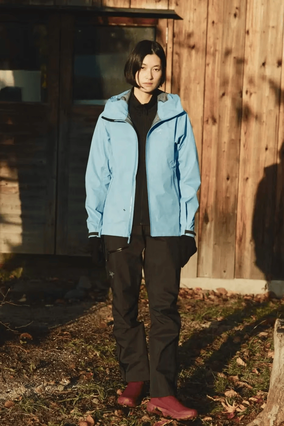 Woman in blue Arc'teryx jacket and black Beams Boy pants in front of a wooden cabin