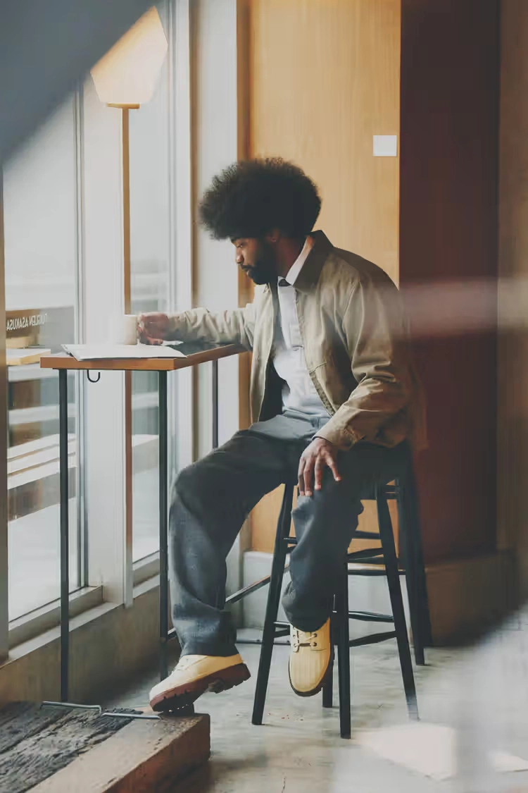 man wearing a khaki jacket and gray sweater, captured next to a blurred window