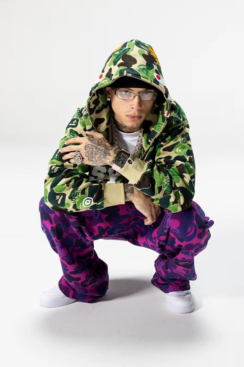 Central Cee avec hoodie Bape camouflage et pantalon violet sur fond blanc