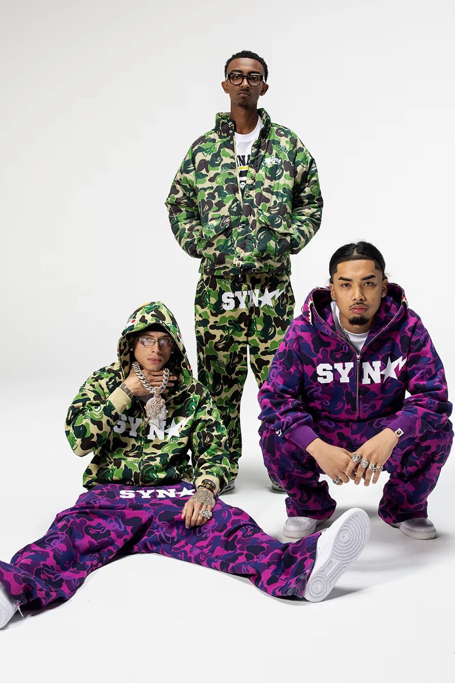 Trois mannequins portant des ensembles BAPE x Spotify SYNA camo vert et violet sur fond blanc