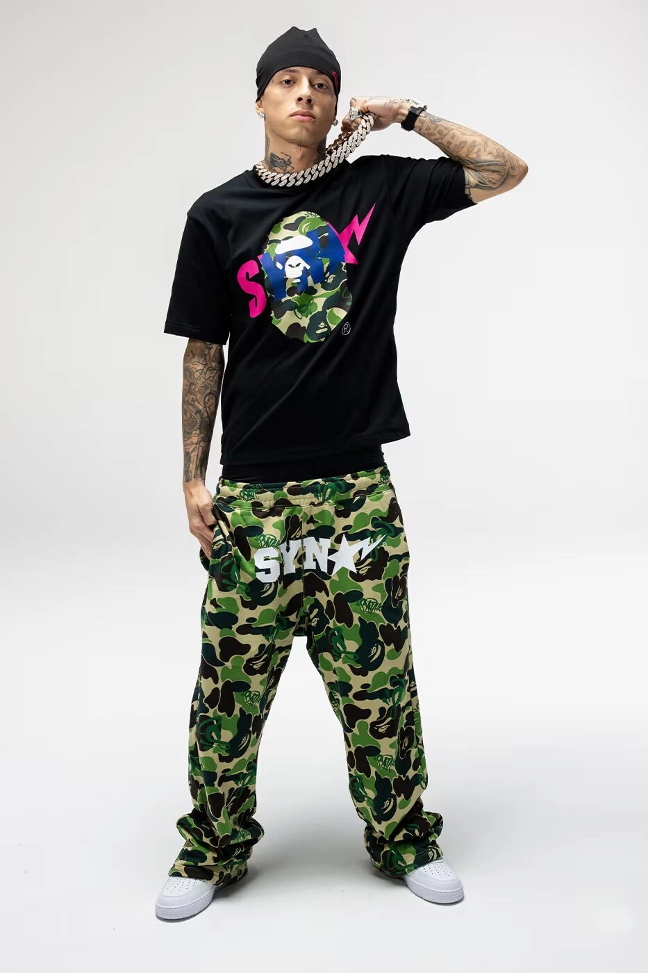 Central Cee portant un t-shirt noir BAPE x Syna et un pantalon camouflage, chaîne autour du cou