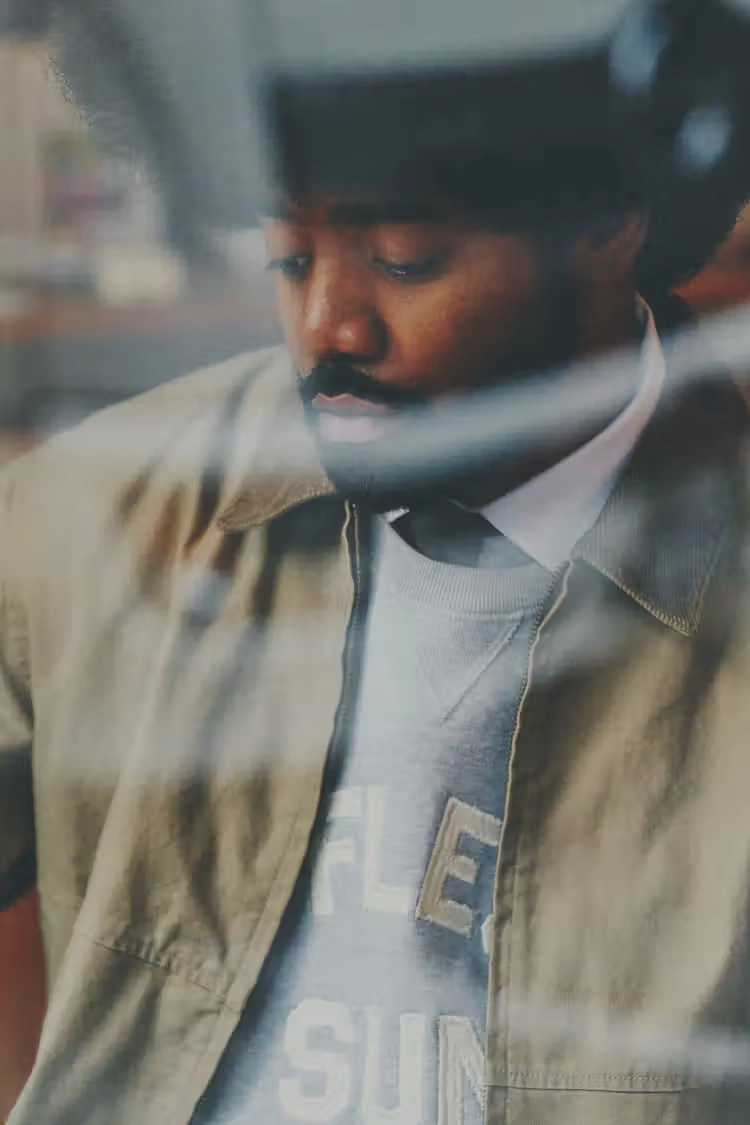 man wearing a khaki jacket and gray sweater, captured through a blurred window