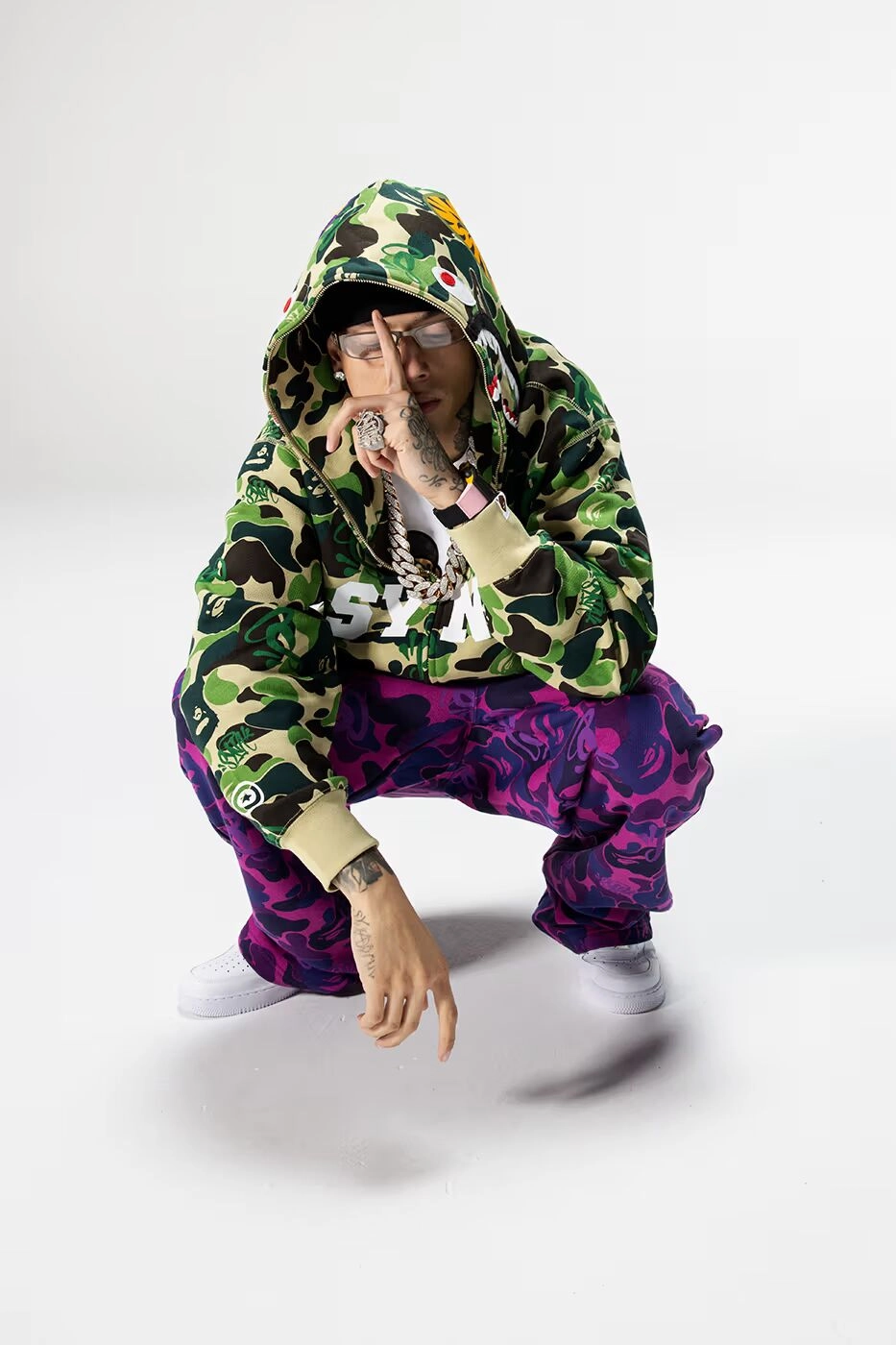 Central Cee portant un ensemble BAPE camouflage vert et violet, accroupie sur fond blanc