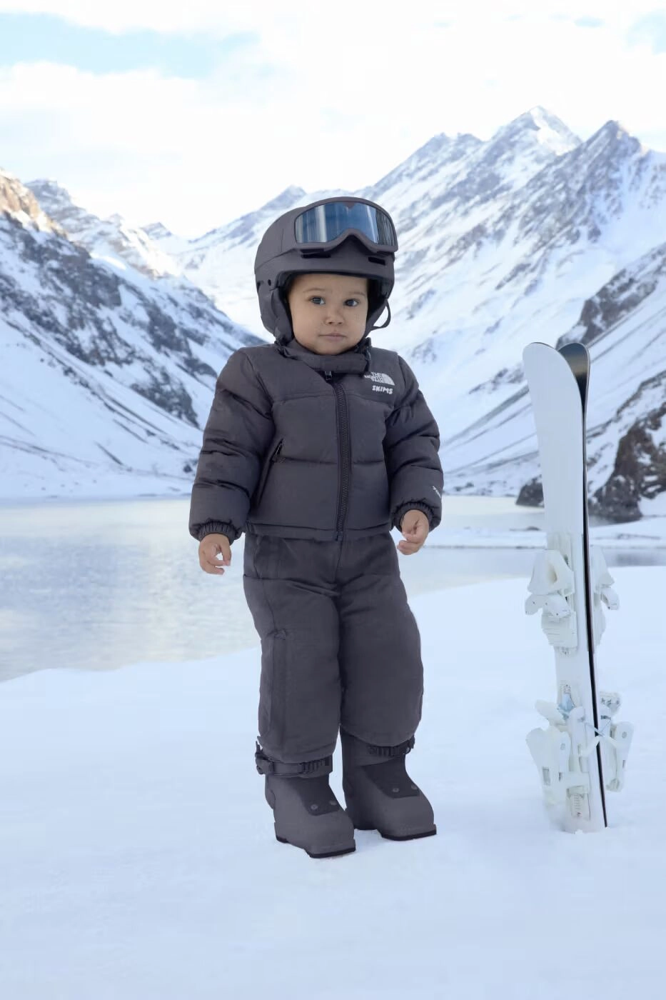 Enfant en combinaison noire SKIMS x The North Face avec casque et skis en montagne enneigée