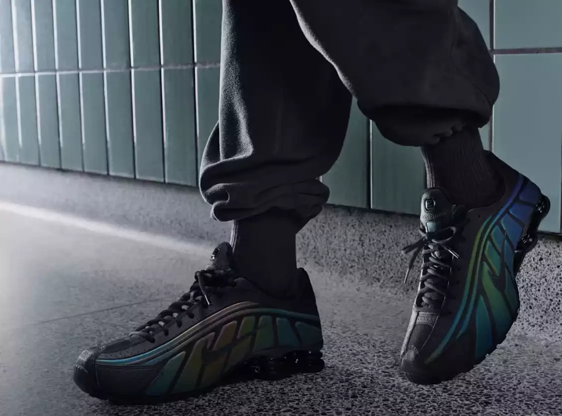 Sneaker Nike Shox noire avec motifs multicolores, portée devant un mur vert